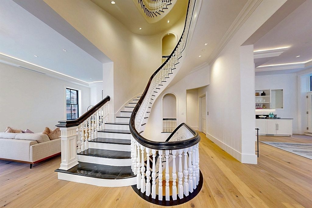 This elegant foyer features a grand, curved staircase with dark-stained treads and a white balustrade that serves as the central architectural focal point. The space is finished with light wood flooring and crisp white walls, creating a bright and airy atmosphere that flows into an adjacent living area and a wet bar. The perspective captures the sweeping curve of the staircase, emphasizing the home's sophisticated and open floor plan.