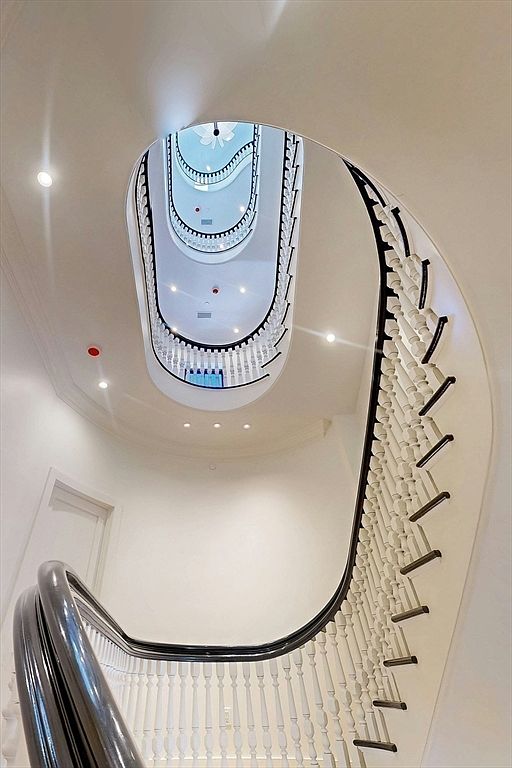 This image captures a dramatic, upward-looking perspective of a grand, multi-story spiral staircase. The design features a dark, polished handrail that curves elegantly along a series of white balusters, creating a striking contrast against the clean, neutral walls. The open central void allows for a clear view of the upper levels, emphasizing the architectural scale and sophisticated flow of the home's interior.