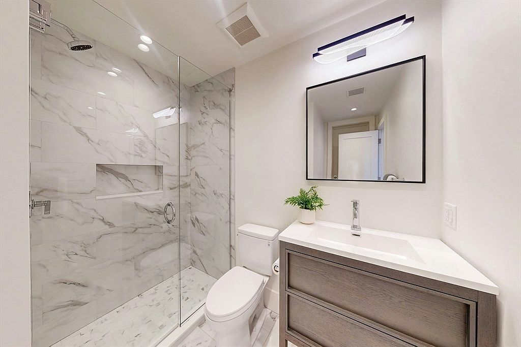 This modern bathroom features a walk-in shower enclosed by a glass panel, showcasing light-toned stone-look wall tiles with a built-in recessed niche. A vanity with a dark wood-toned cabinet and a white countertop sits beside a white toilet, complemented by a rectangular mirror and a sleek light fixture above. The space is bright and clean, offering a contemporary aesthetic suitable for a guest or secondary bathroom.