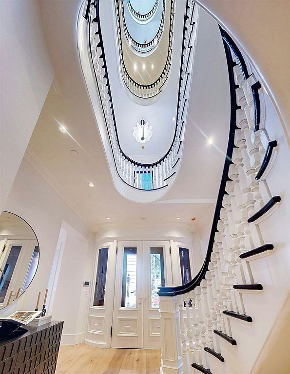 This grand entryway features a dramatic, multi-story spiral staircase with white balusters and a dark handrail, creating a striking architectural focal point. The space is bright and airy, with light wood flooring and a set of double doors leading to the exterior. A modern, sculptural light fixture hangs from the ceiling, drawing the eye upward through the open stairwell.