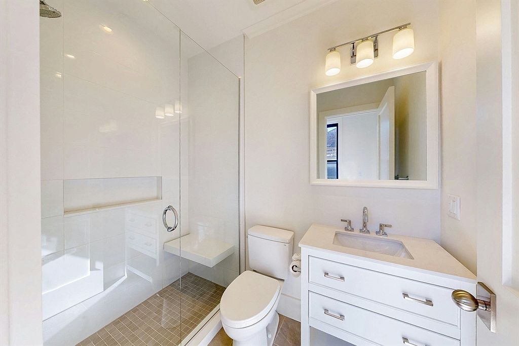 This bright, compact bathroom features a walk-in shower with a glass panel, a built-in bench, and a tiled floor. A white vanity with a stone countertop and a rectangular sink sits beside a standard toilet, all illuminated by a three-light fixture above the mirror. The space is finished in a clean, neutral palette, creating a functional and modern aesthetic.