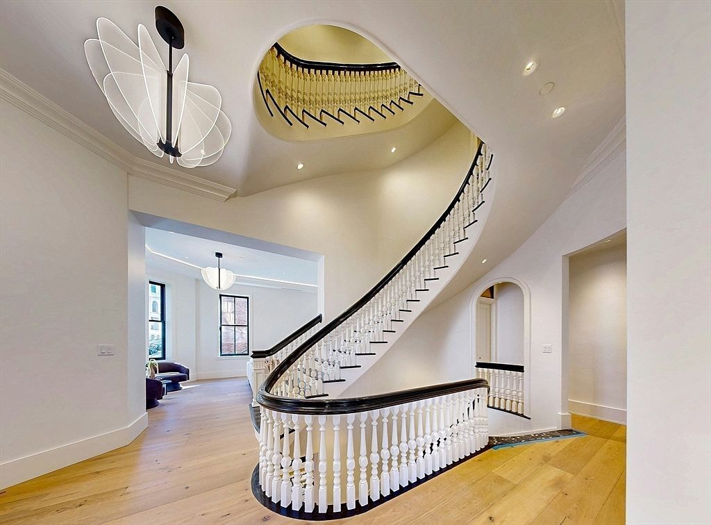 This grand foyer features a striking curved staircase with a dark wood handrail and white balusters, serving as the central architectural focal point. The space is illuminated by a unique, sculptural modern chandelier and recessed lighting, casting a warm glow over the light wood flooring. The open floor plan allows for a clear view into an adjacent living area, creating an airy and sophisticated atmosphere.