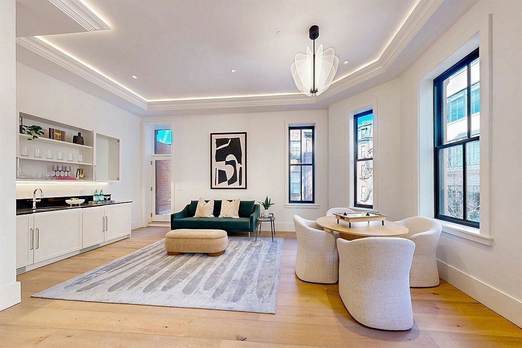 This bright and airy living space features a contemporary design with light wood flooring and a neutral color palette. The room includes a wet bar area with white cabinetry, a plush green sofa paired with a beige ottoman, and a circular dining table surrounded by four upholstered chairs. Large windows with black frames allow for ample natural light, while a unique modern chandelier serves as a focal point above the seating area.