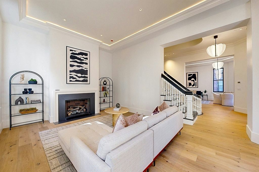 This bright and airy living room features light wood flooring, a neutral-toned sofa, and a central fireplace with a dark, minimalist surround. The space is accented by two arched metal shelving units and abstract wall art, with a staircase leading to an adjacent room visible in the background. The perspective is from the living area looking toward the staircase, highlighting the open-concept flow and modern aesthetic.