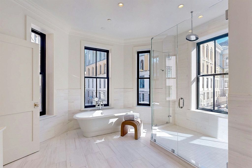 This bright and airy primary bathroom features a freestanding soaking tub positioned beneath a large window, complemented by a separate glass-enclosed shower. The room is finished with light-toned stone tile flooring and walls, creating a clean, spa-like aesthetic. The perspective captures the entire space, highlighting the natural light streaming in through multiple tall windows that offer views of the surrounding urban architecture.