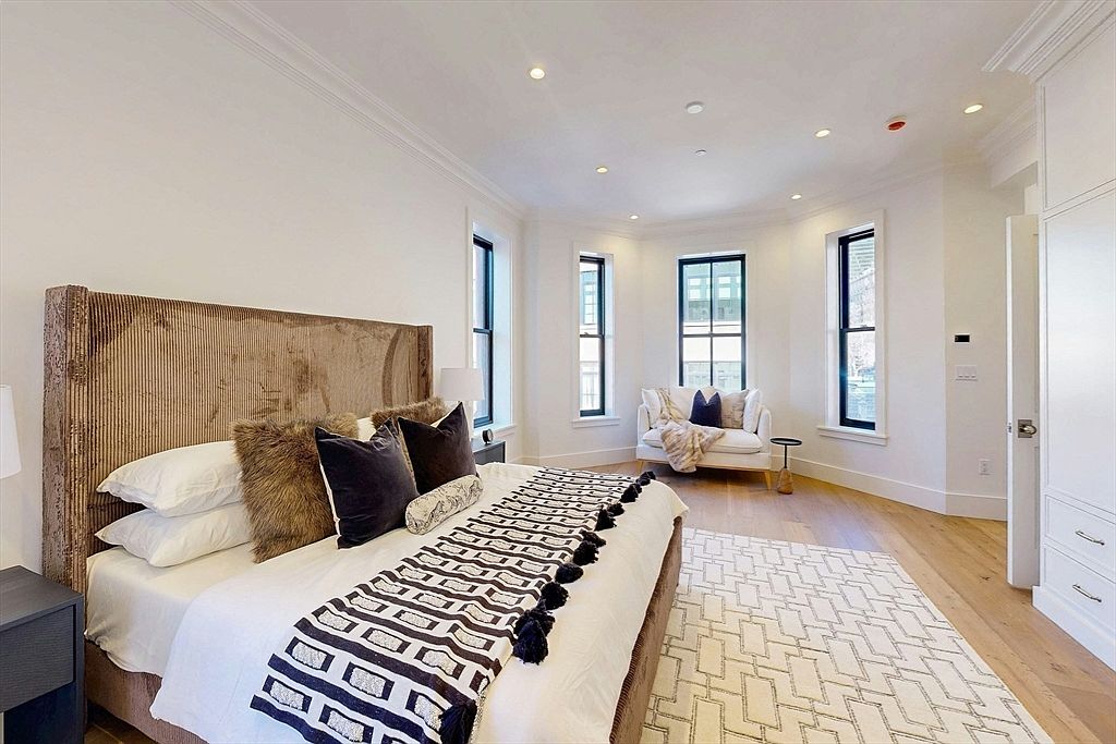 This bright and airy primary bedroom features a large bed with a textured, tall headboard and a patterned throw blanket. The room is anchored by a geometric-patterned area rug over light wood flooring and includes a cozy seating area nestled in a bay window nook. The space is illuminated by recessed lighting and natural light streaming through multiple windows, creating a clean and sophisticated atmosphere.