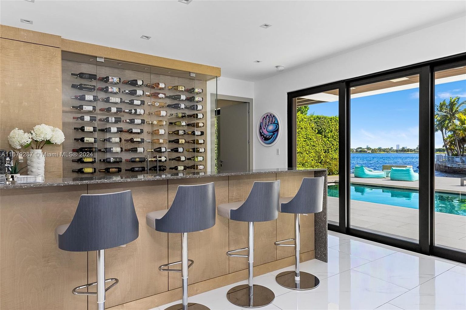 This interior shot showcases a stylish wine cellar and bar area, complete with a large wine rack filled with bottles. Four modern bar stools line the counter, and sliding glass doors offer a view of the pool and waterfront. The space is bright and airy, with white tile flooring and a contemporary design.