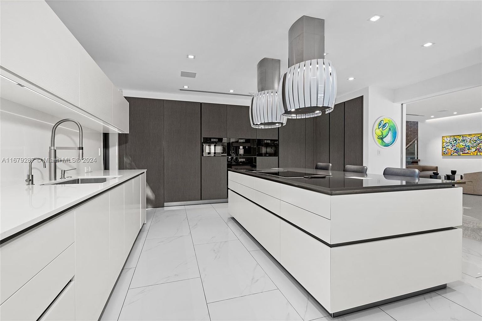 This is a modern kitchen with a sleek design. It features white cabinetry, a dark countertop island with seating, and unique pendant lighting. The kitchen also includes built-in appliances and a minimalist aesthetic, creating a clean and sophisticated space.