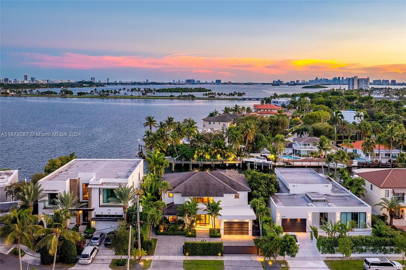This aerial view showcases a collection of luxurious waterfront homes at sunset. The properties feature modern architectural designs, lush landscaping with palm trees, and well-manicured lawns. The scene is set against a backdrop of a calm bay and a distant city skyline, creating an impression of upscale coastal living.