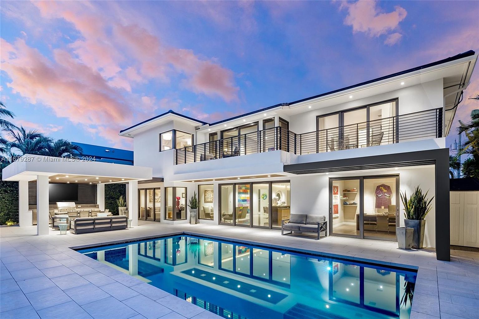 This is a rear exterior view of a modern two-story home featuring a swimming pool and an outdoor kitchen area. The house is painted white with black accents, including balcony railings and window frames. The pool reflects the house and the colorful sky, and the outdoor kitchen is equipped with a grill and seating area, creating an inviting space for relaxation and entertainment.