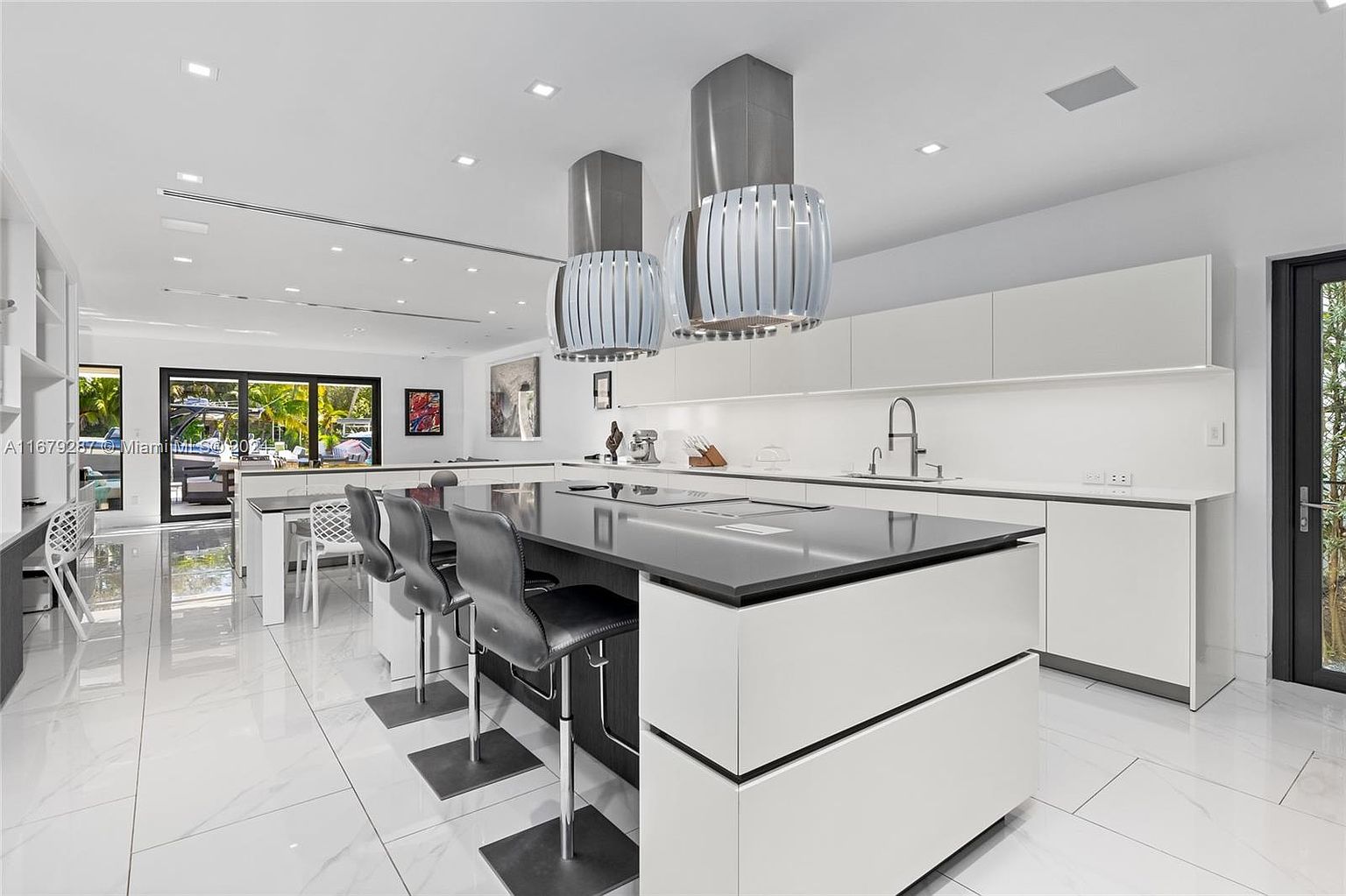 This is a modern kitchen with a large island featuring a black countertop and white base, complemented by sleek gray bar stools. The kitchen is equipped with stainless steel appliances and unique pendant lighting fixtures, set against a backdrop of white cabinetry and a bright, open floor plan with large windows offering natural light.