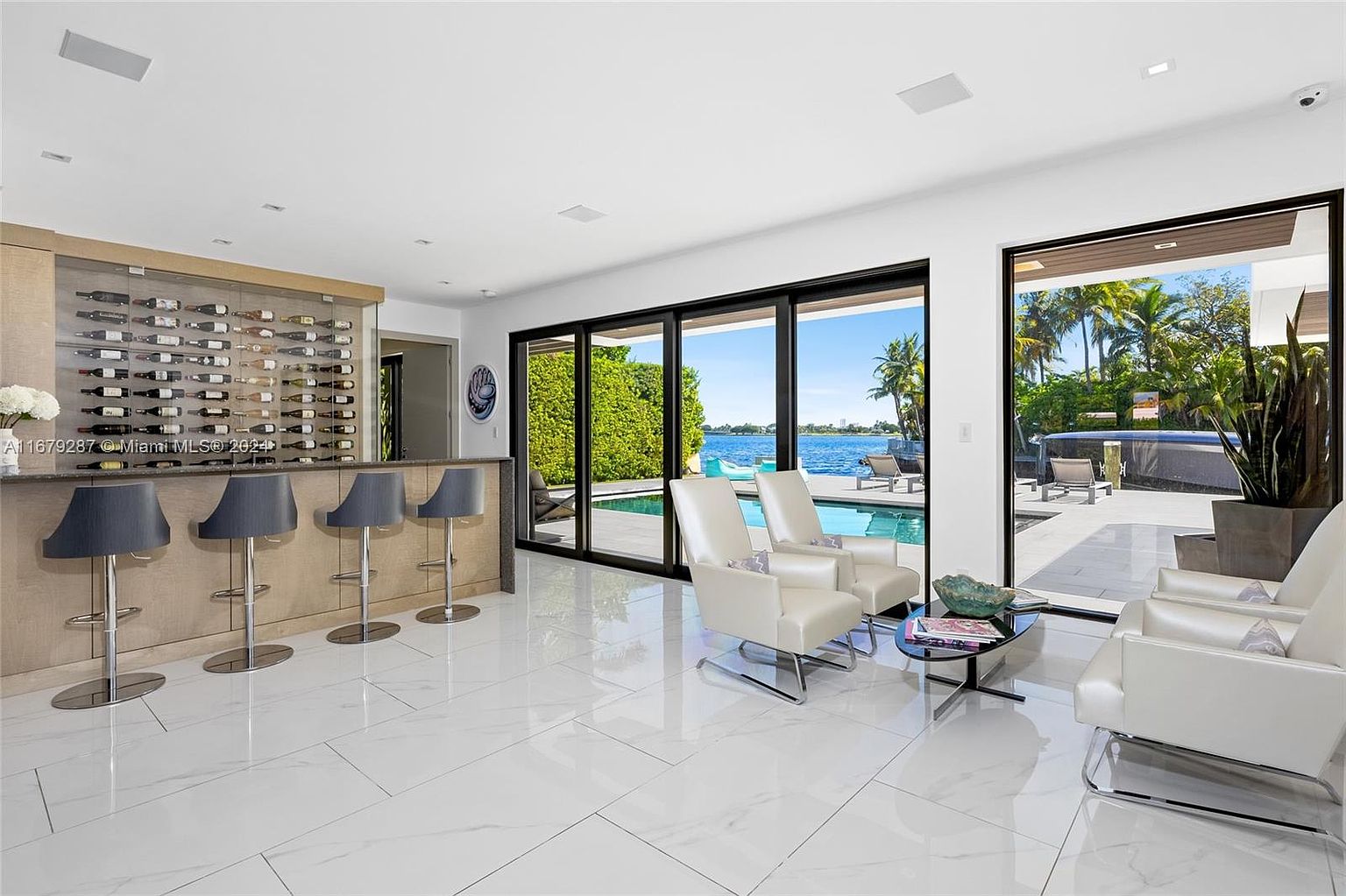 This interior shot showcases a luxurious living area with a modern aesthetic. The room features a wine rack and bar area, sleek white tile flooring, and comfortable white armchairs. Large sliding glass doors offer a seamless transition to an outdoor pool and patio area, providing stunning waterfront views and an abundance of natural light.