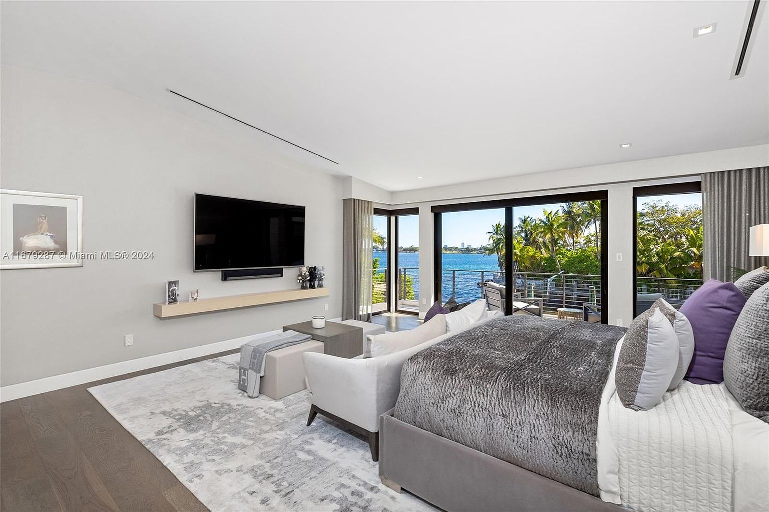 This is a luxurious primary bedroom featuring a large bed with a plush gray comforter and decorative pillows. A flat-screen TV is mounted on the wall above a floating shelf, and sliding glass doors offer stunning water views and access to a balcony. The room is decorated in a modern style with a neutral color palette and a stylish area rug.