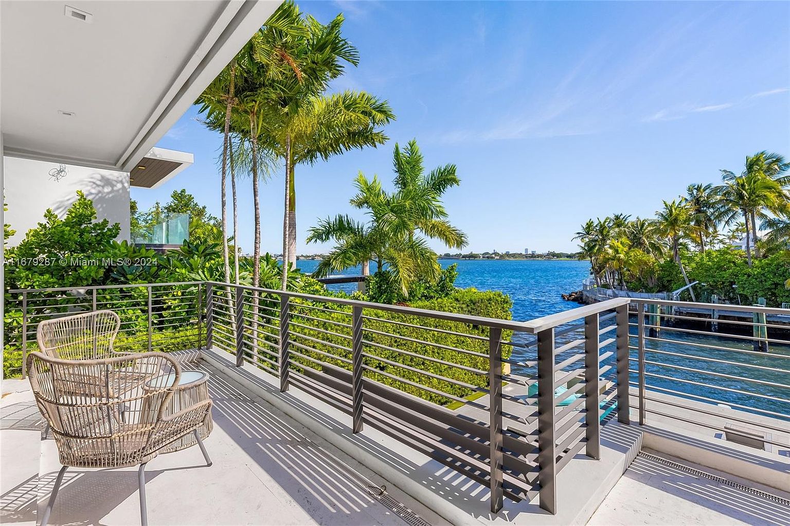 This image showcases a luxurious balcony with a stunning waterfront view. The balcony features modern metal railings and comfortable wicker furniture, creating an inviting outdoor living space. Lush tropical foliage and palm trees enhance the serene atmosphere, making it an ideal spot for relaxation and enjoying the beautiful scenery.