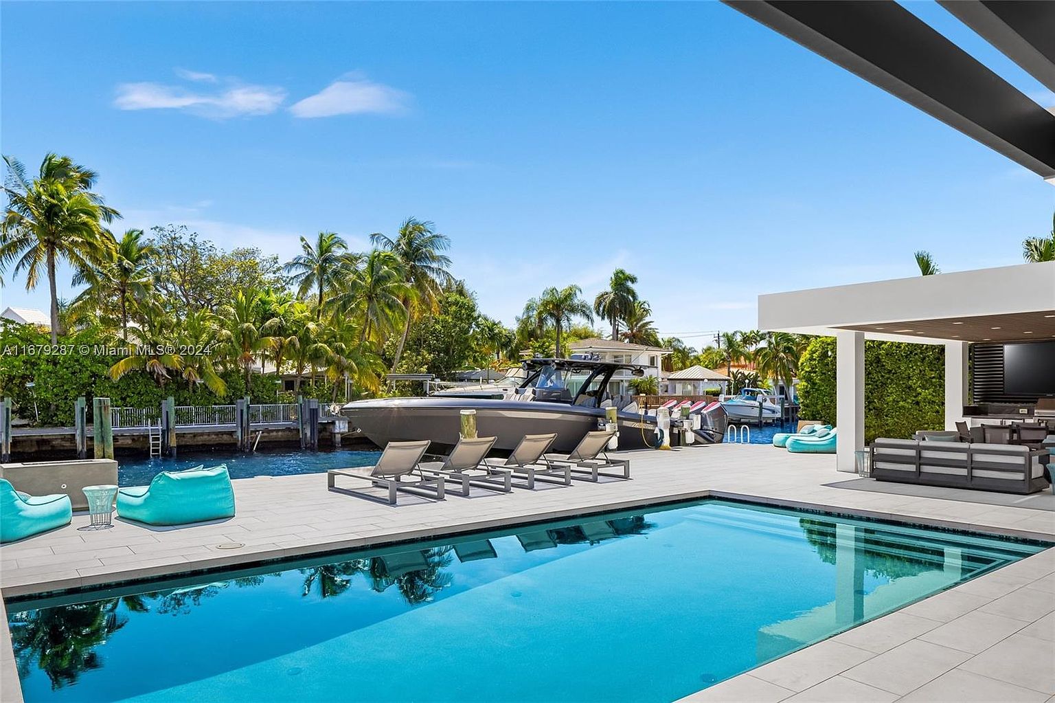 This image showcases a luxurious outdoor space featuring a pristine swimming pool with clear blue water, surrounded by a modern patio area. The scene includes stylish lounge chairs, a boat docked nearby, and a covered outdoor living area with comfortable seating, suggesting a high-end waterfront property designed for relaxation and entertainment. The overall impression is one of sophistication and leisure.