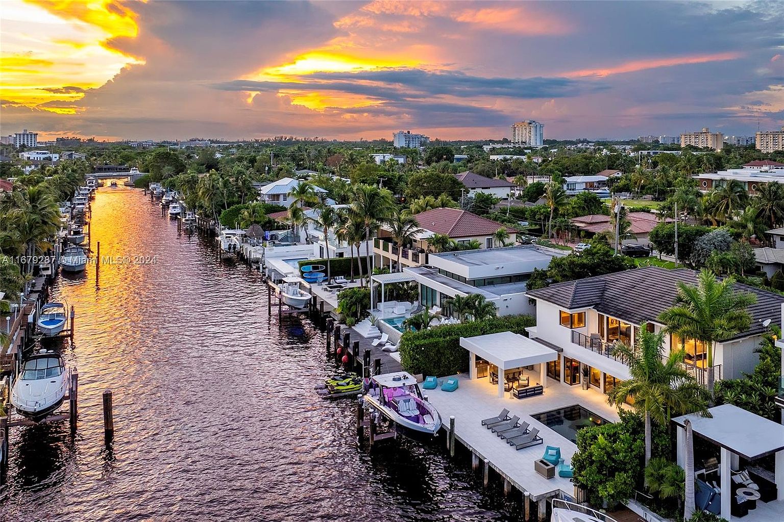 This aerial view showcases a luxurious waterfront property with a private dock and multiple boats. The modern architecture of the homes is complemented by lush landscaping and a serene canal setting. The image captures a vibrant sunset, enhancing the overall appeal and highlighting the upscale neighborhood.