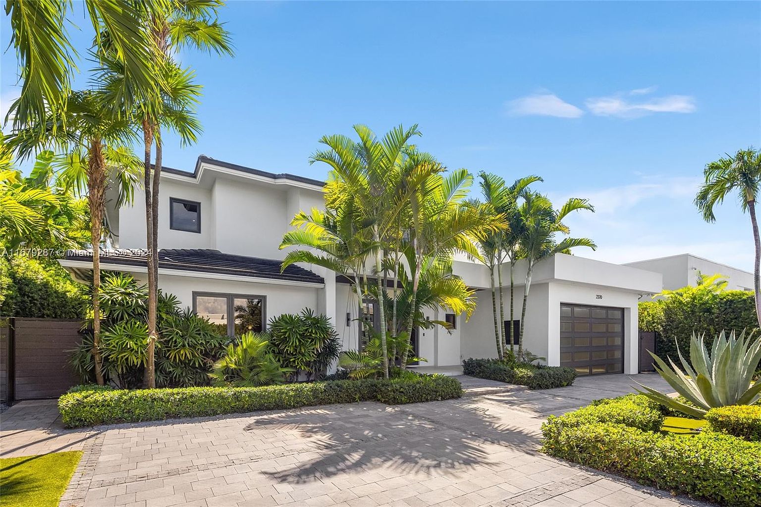 This is a front exterior view of a modern two-story home with a clean, white facade and a dark roof. The property features lush tropical landscaping, including palm trees and manicured hedges, enhancing its curb appeal. A paved driveway leads to a garage, and the overall impression is one of contemporary elegance and privacy.