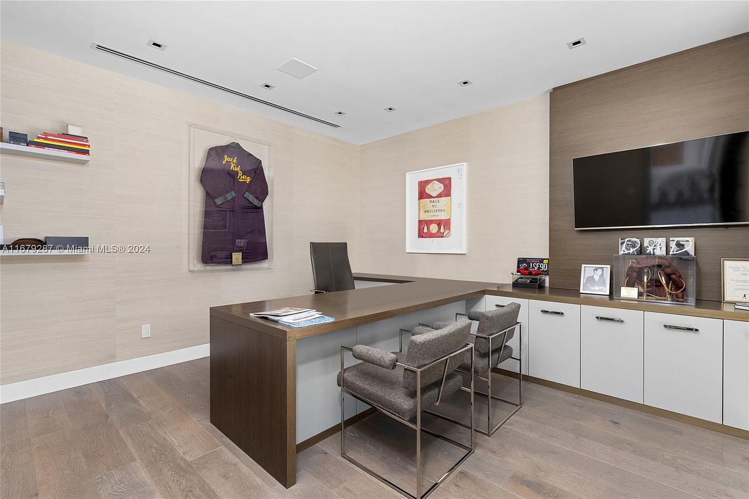This is a well-appointed home office featuring a large, L-shaped desk with a dark wood finish and light gray accents. Two modern chairs with metal frames are positioned in front of the desk. The room is decorated with framed artwork, a flat-screen television, and various decorative items, creating a sophisticated and functional workspace.