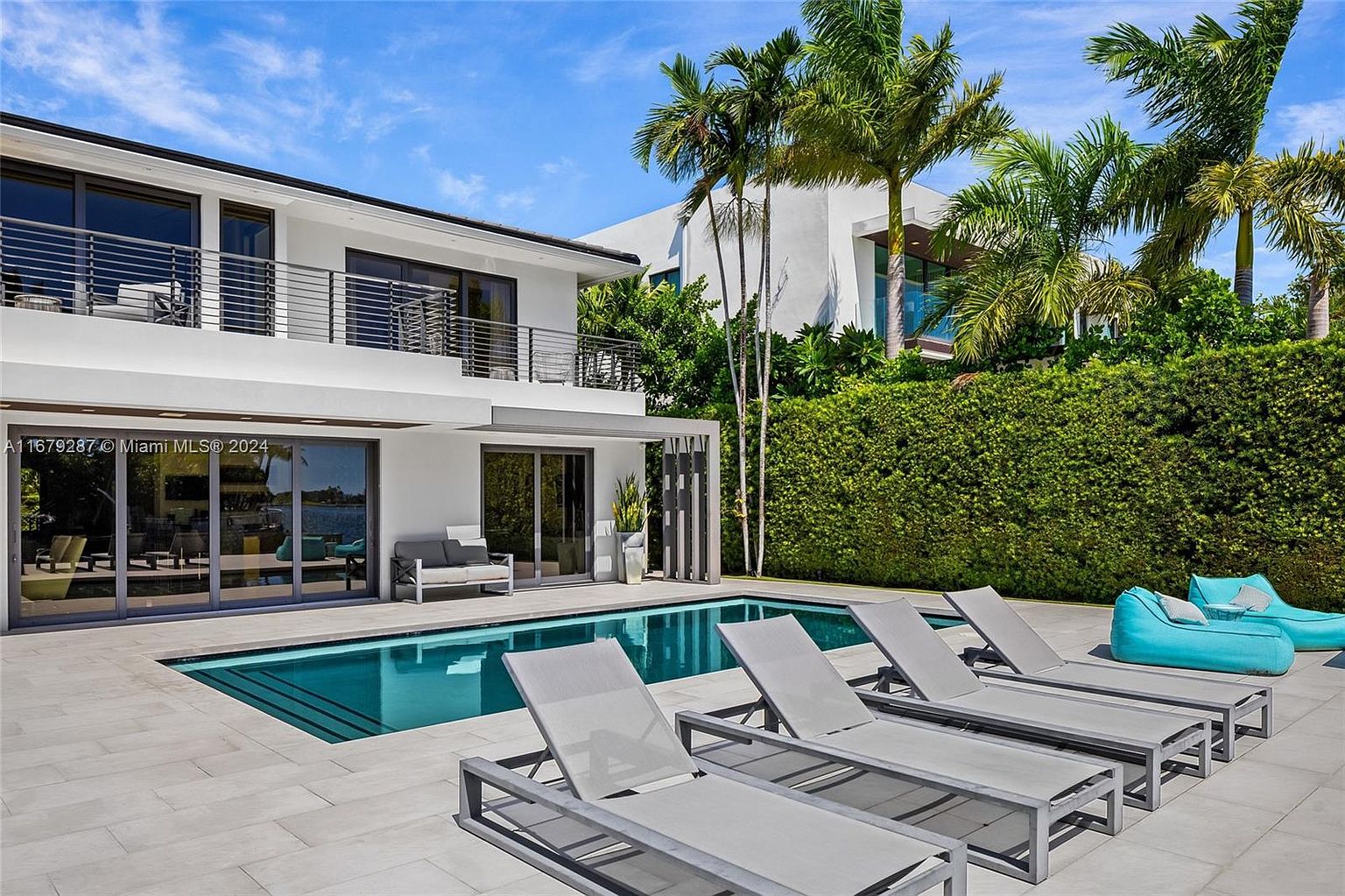 This image showcases a luxurious backyard featuring a modern house with a pool and lounge area. The pool is surrounded by sleek gray lounge chairs and a turquoise beanbag, creating a relaxing atmosphere. Lush greenery and palm trees add to the tropical feel, making it an ideal outdoor space for entertaining or unwinding.