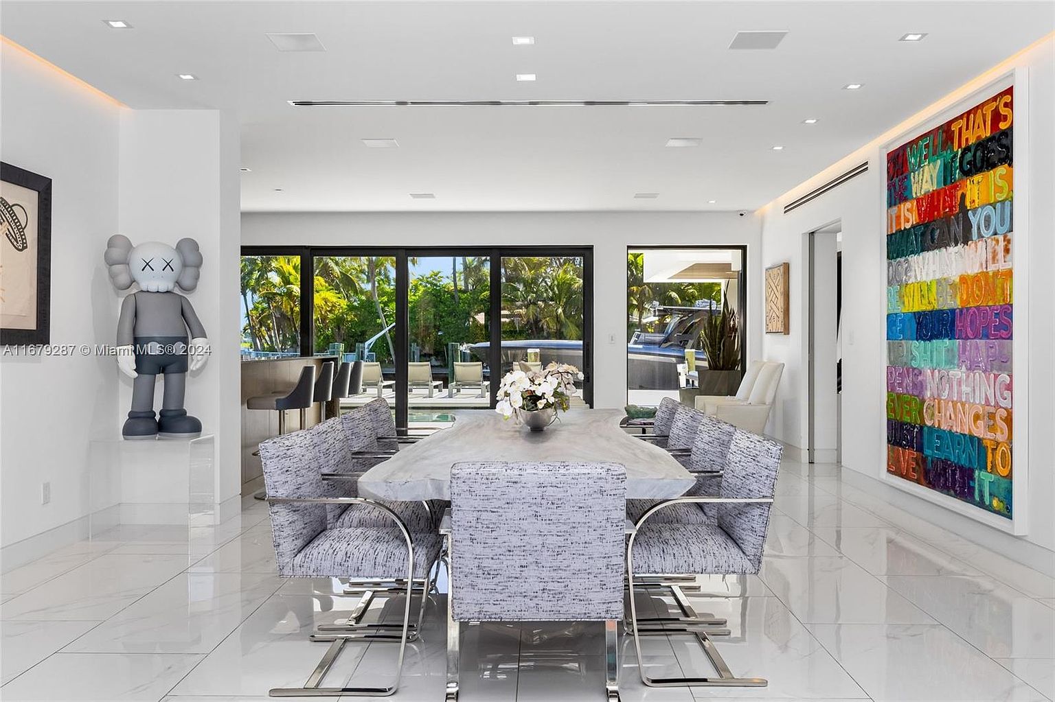 This interior shot showcases a modern dining room with a large, uniquely shaped dining table surrounded by upholstered chairs with chrome frames. The room features white marble flooring, contemporary artwork, and floor-to-ceiling windows that offer a view of the outdoor pool area and lush greenery. A KAWS sculpture adds a touch of modern art.