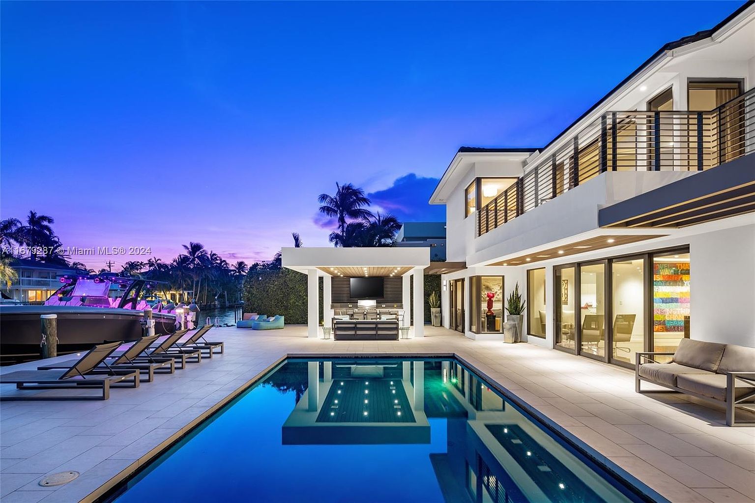 This is a stunning exterior shot of a modern luxury home at dusk, showcasing a beautiful pool and outdoor entertainment area. The pool's reflective surface mirrors the architectural details of the house and the outdoor kitchen/bar area, creating a symmetrical and visually appealing composition. Lounge chairs are placed near the pool, and a boat is docked nearby, suggesting a waterfront property with upscale amenities.