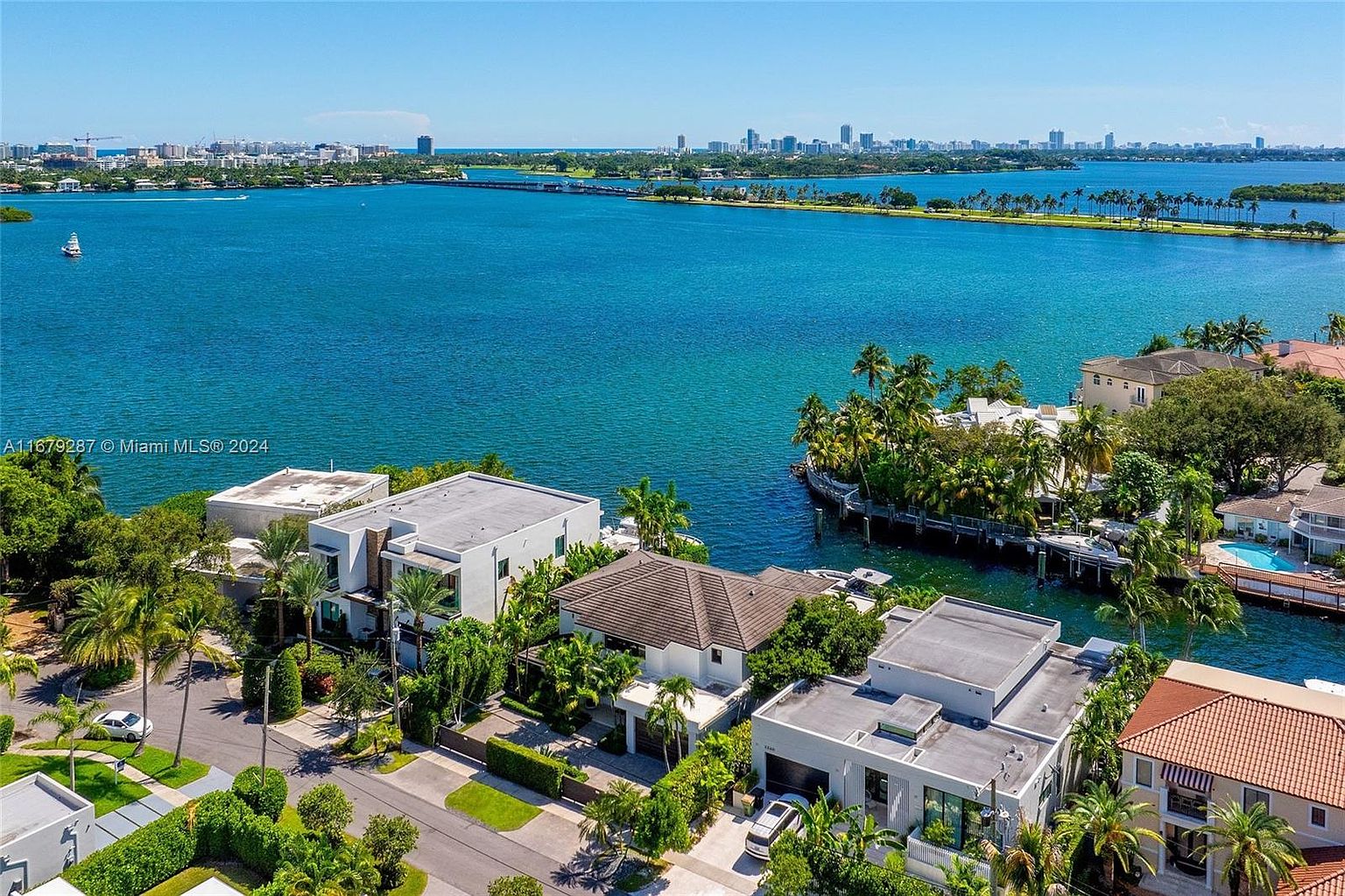This aerial view showcases a collection of luxury waterfront homes, each with unique architectural designs and lush landscaping. The properties are situated along canals and the bay, offering direct water access and stunning views of the city skyline in the distance. The scene conveys a sense of exclusivity and high-end living in a tropical paradise.