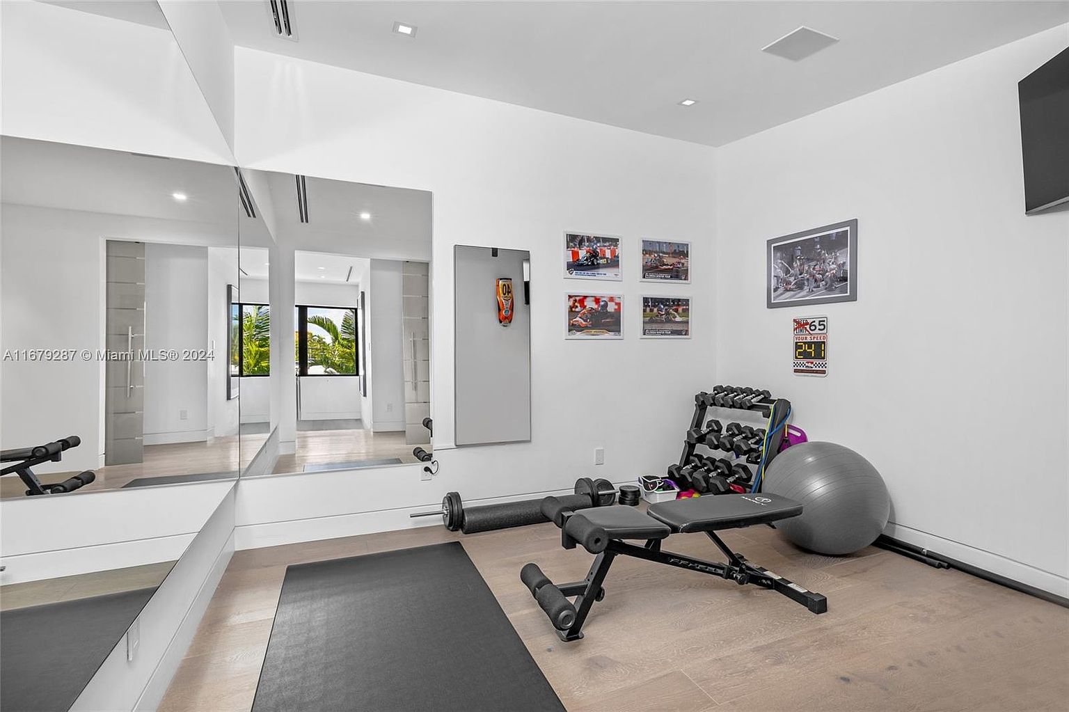 This is a well-equipped home gym featuring a weight bench, dumbbell rack, exercise ball, and a large mirror that visually expands the space. The room has light hardwood flooring and white walls adorned with framed pictures, creating a clean and motivating workout environment. The presence of a black exercise mat suggests a dedicated area for floor exercises.