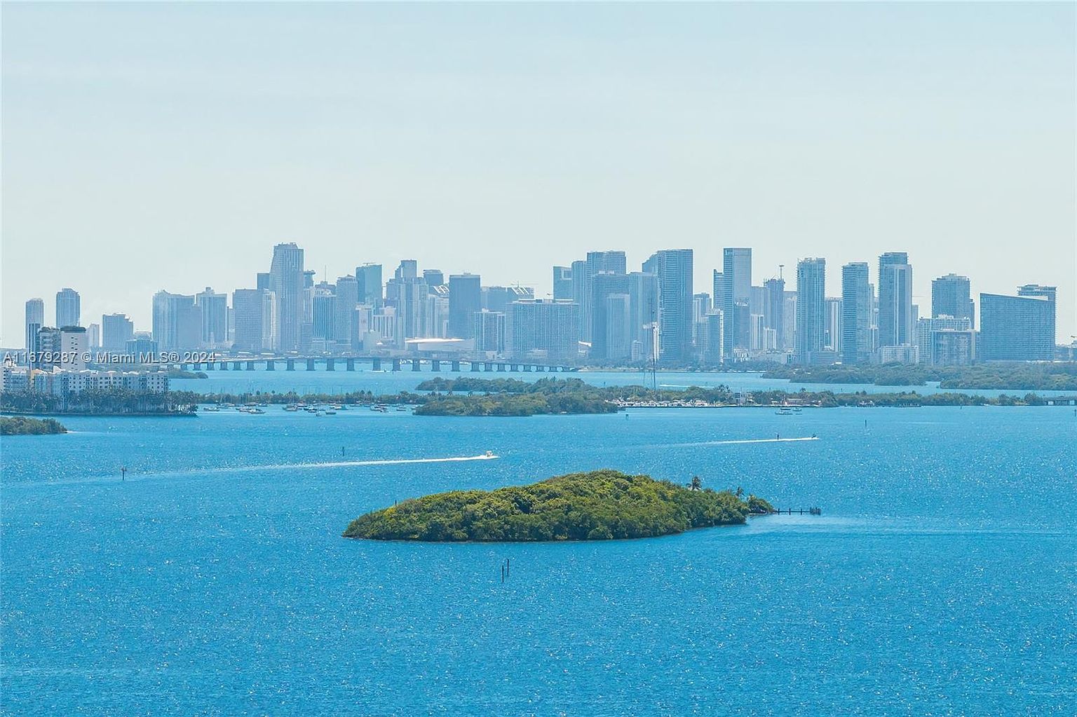 This aerial view showcases a stunning cityscape backdrop with a tranquil island in the foreground. The turquoise water adds a vibrant contrast to the lush greenery of the island and the towering buildings in the distance. The scene evokes a sense of serenity and exclusivity, highlighting the desirable coastal location.