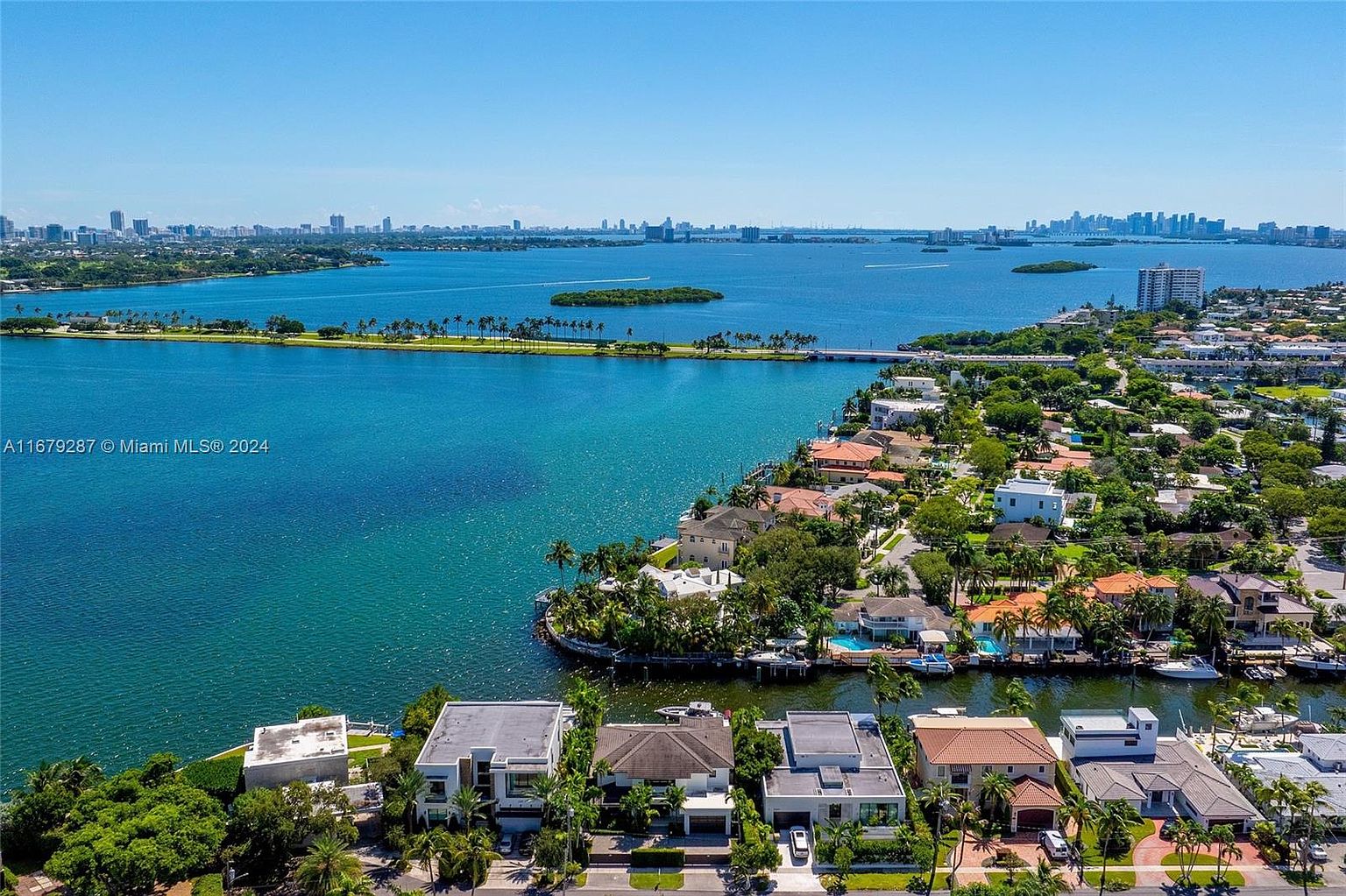 This aerial shot showcases a luxurious waterfront property with stunning views of the bay and city skyline. The homes feature well-manicured lawns, private docks, and some with pools, creating an exclusive and desirable neighborhood. The turquoise waters and clear blue sky enhance the overall appeal, highlighting the prime location and upscale lifestyle.