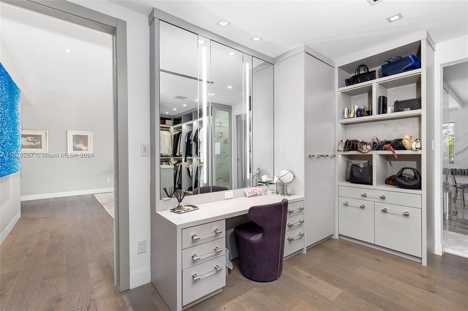 This is a well-organized walk-in closet featuring a vanity area with a mirrored wall and a built-in storage unit with open shelving and drawers. The closet is illuminated by recessed lighting and features a purple upholstered chair. The overall design is modern and luxurious, providing ample storage and a dedicated space for dressing and grooming.