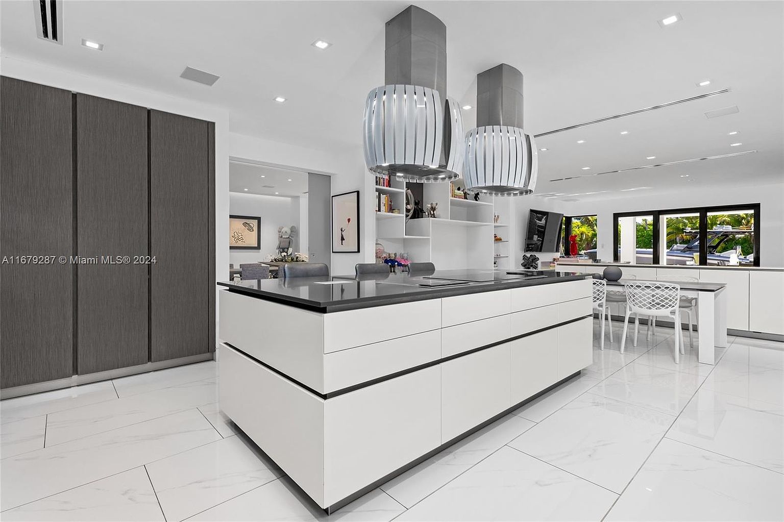 This is a modern kitchen featuring a large white island with a black countertop, complemented by unique pendant lighting fixtures. The kitchen boasts sleek cabinetry and an open layout, leading to a dining area with white chairs and a view of the outdoors through large windows. The overall design is minimalist and sophisticated, emphasizing clean lines and a bright, airy atmosphere.