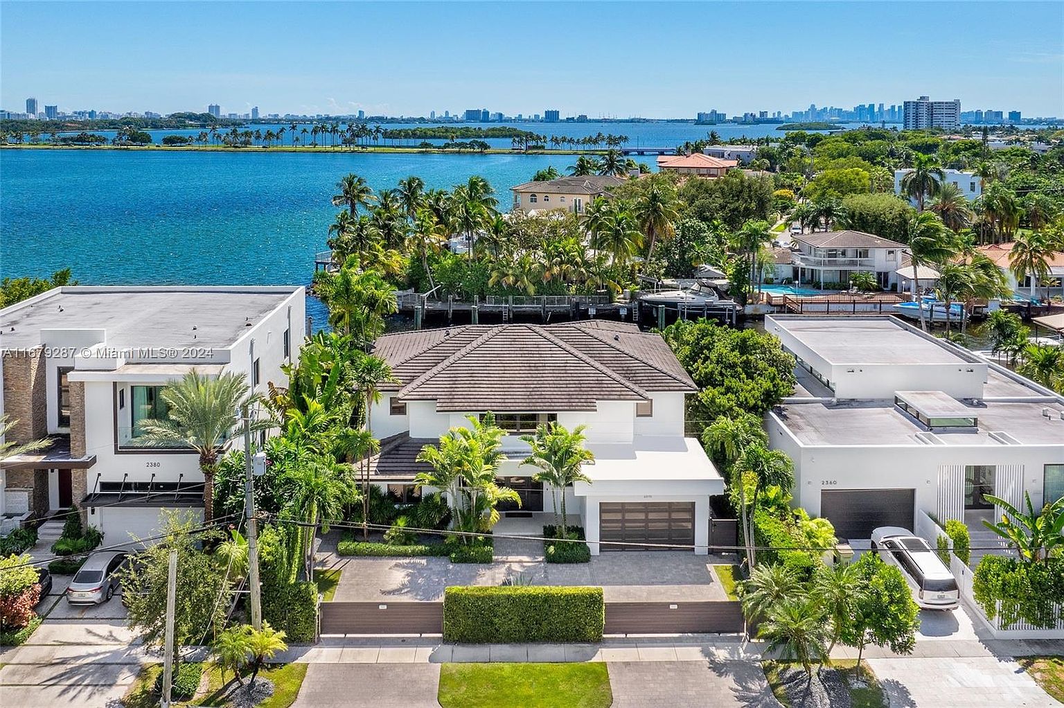 This aerial shot showcases a luxurious waterfront property with a modern architectural design. The house features a brown tiled roof, a white exterior, and lush landscaping with palm trees. A gated entrance provides privacy, and the view extends to the bay and the city skyline in the distance, creating an impression of exclusivity and high-end living.