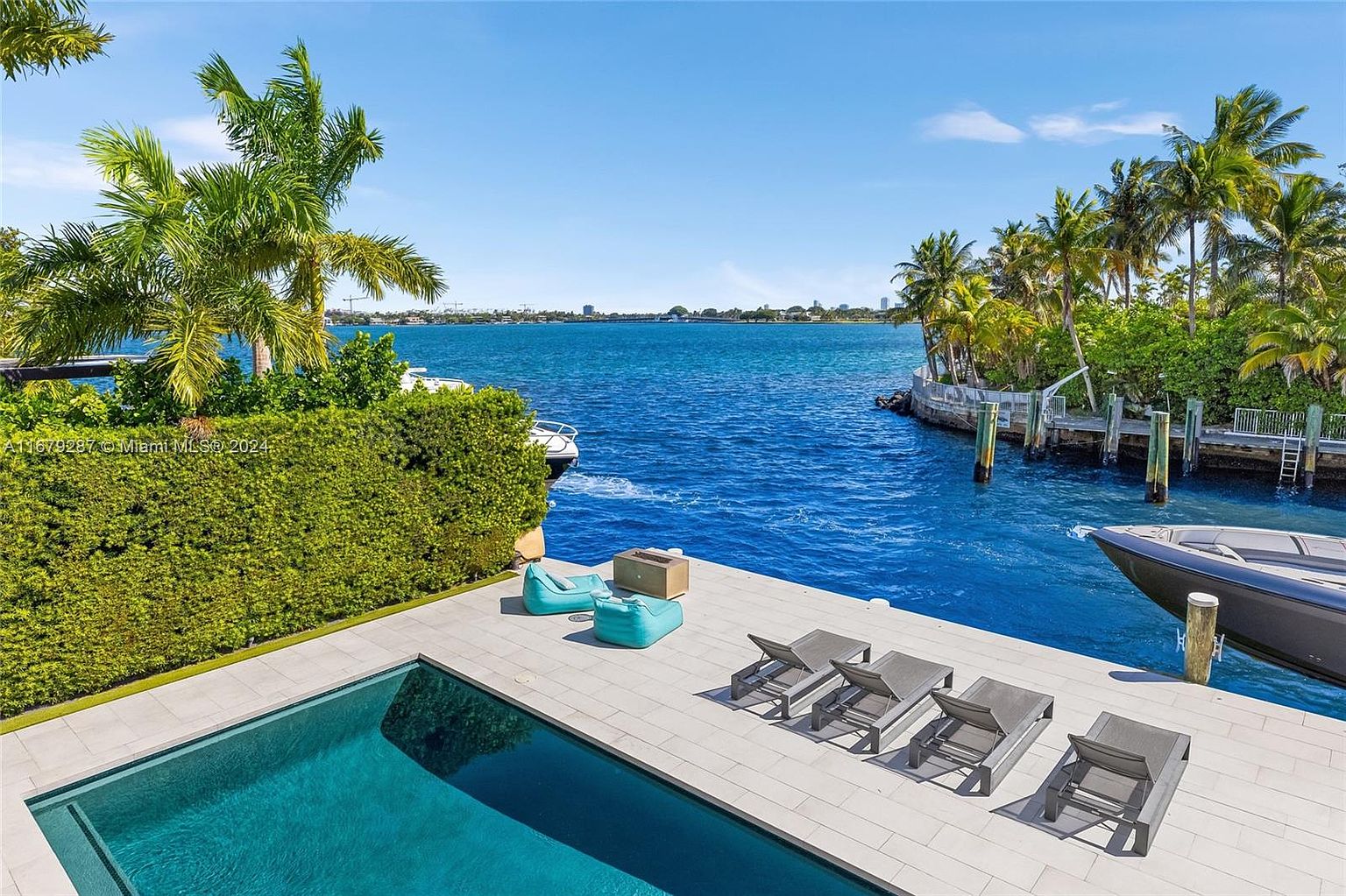 This exterior shot showcases a luxurious pool area with modern design elements. The pool is surrounded by a sleek, light-colored patio featuring lounge chairs and comfortable seating. The property extends to the waterfront, offering stunning views of the ocean and a private dock with a boat, creating an inviting and upscale outdoor living space.