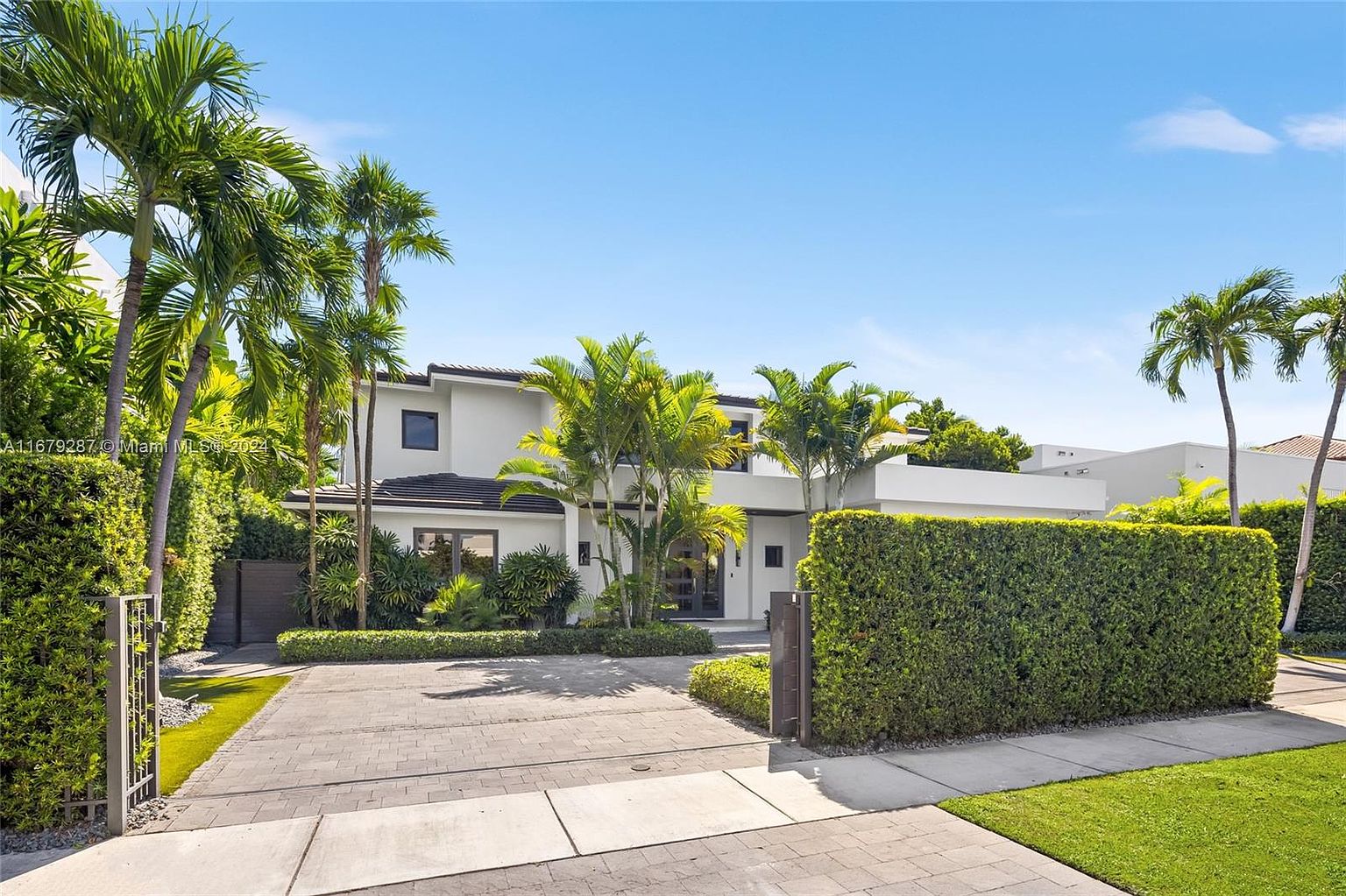 This is a front exterior view of a modern, two-story home with a white facade and a dark tile roof. The property is surrounded by lush landscaping, including mature palm trees and manicured hedges, providing privacy and curb appeal. A paved driveway leads to the entrance, enhancing the overall aesthetic and suggesting a well-maintained and luxurious residence.