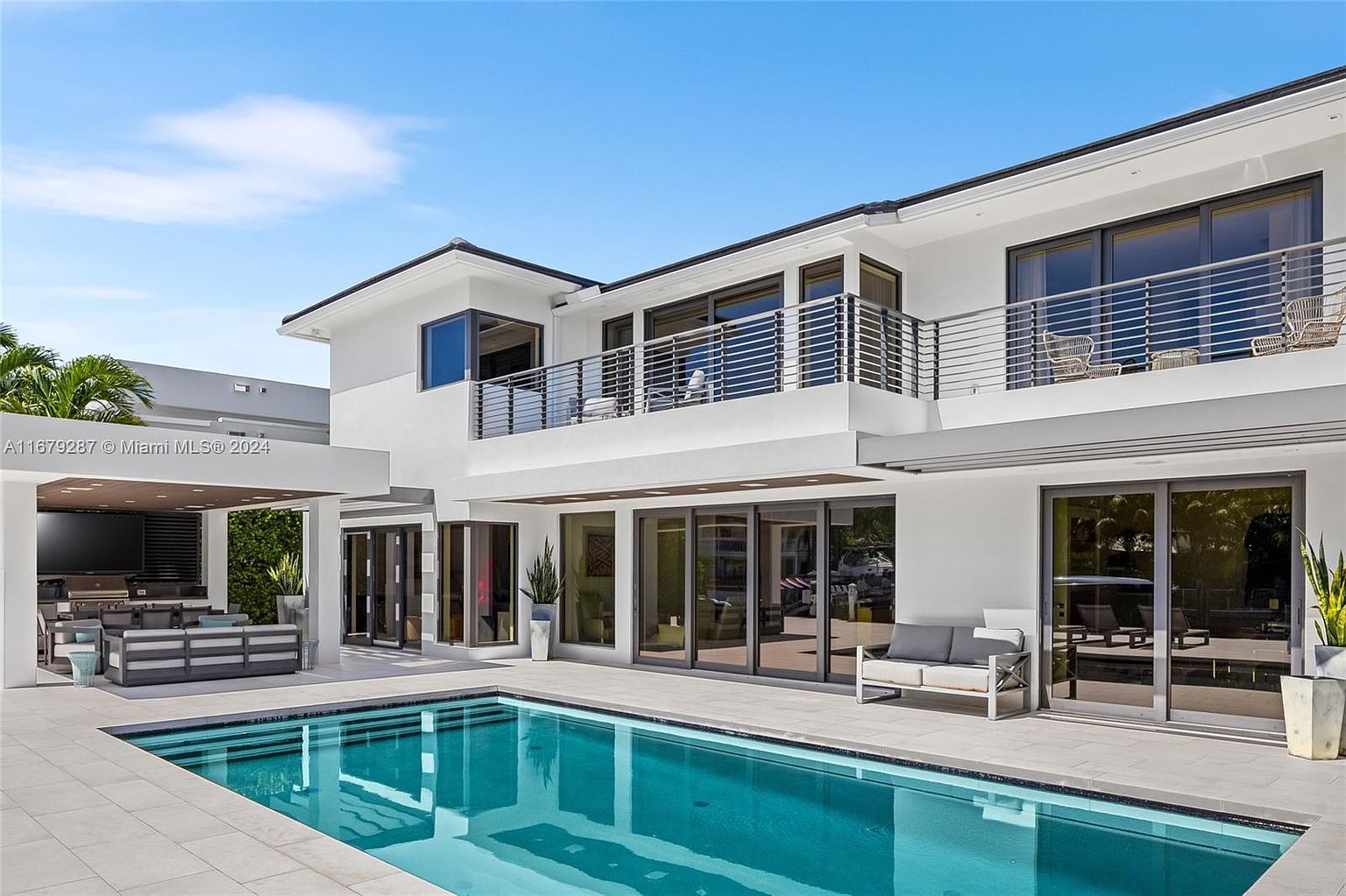 This image showcases a modern home's exterior with a focus on the pool area. The pool features clear blue water and is surrounded by light-colored tiling. The house has a sleek, white facade with large windows and balconies, creating a luxurious and inviting outdoor living space.