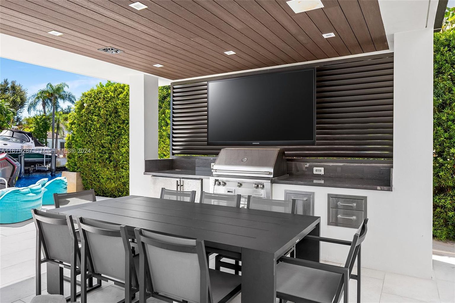This image showcases an outdoor patio area designed for entertainment. It features a built-in grill station with a stainless steel grill and countertop space, complemented by a large flat-screen television mounted above. A modern dining table with chairs is positioned in front, creating a comfortable space for outdoor dining and relaxation.