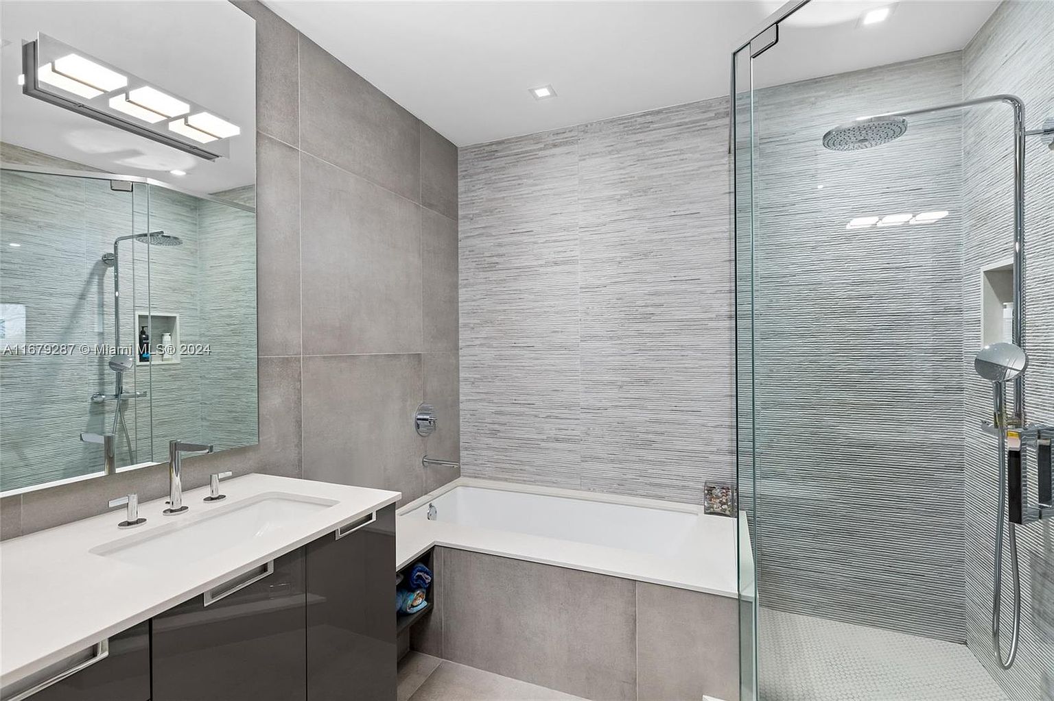 This is a modern primary bathroom featuring a sleek vanity with a white countertop and dark gray cabinetry. A large mirror reflects the shower area, which has a glass enclosure and textured gray tiles. The bathtub is integrated into the shower area, creating a spa-like atmosphere.