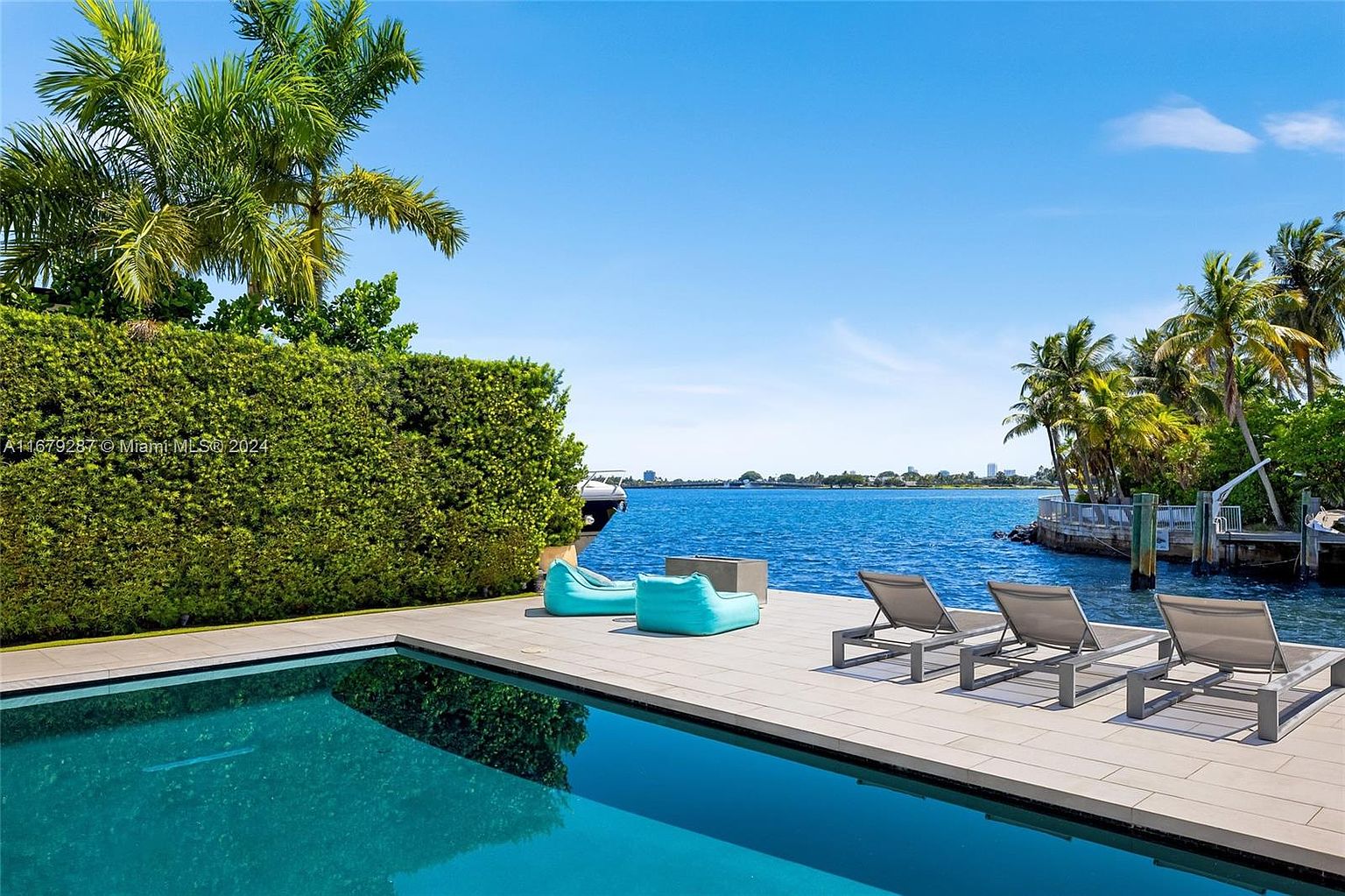 This image showcases a luxurious outdoor pool area with a stunning waterfront view. The pool's turquoise water reflects the clear blue sky, while modern lounge chairs and beanbag chairs are arranged on the tiled deck. Lush greenery provides privacy, and palm trees add a tropical touch, creating an inviting and serene atmosphere.