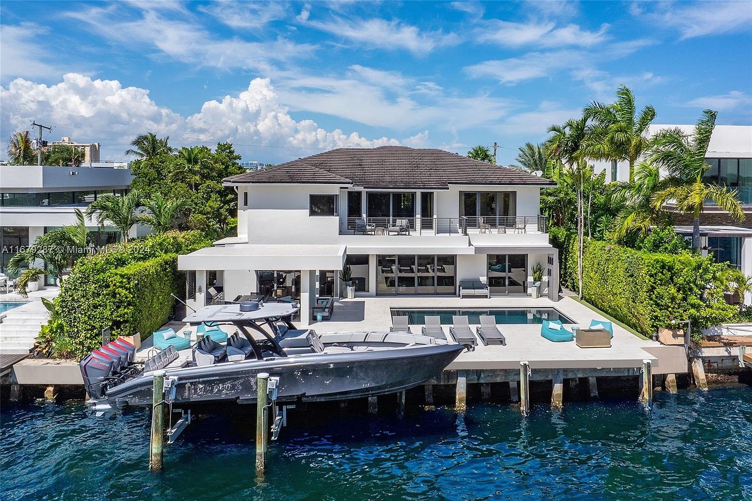 This image showcases the rear exterior of a modern waterfront home. The property features a multi-level deck with lounge chairs and a pool, seamlessly blending indoor and outdoor living. A boat is docked at the property, emphasizing the waterfront lifestyle.
