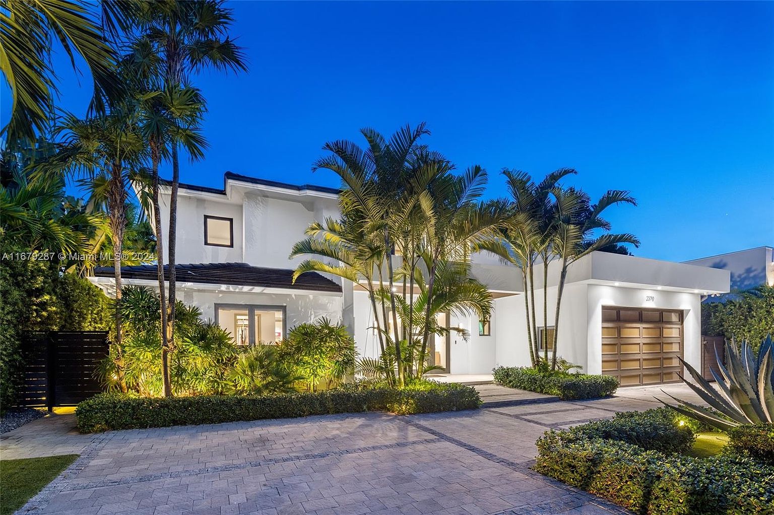 This is a front exterior view of a modern two-story home at dusk. The house features a white facade, a dark roof, and a contemporary garage with a wooden door. Lush landscaping with palm trees and manicured bushes surrounds the property, enhancing its curb appeal and creating a welcoming atmosphere.