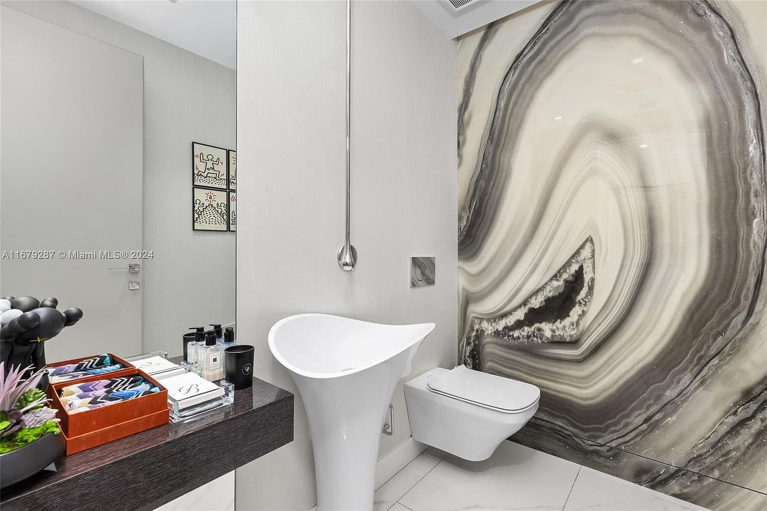 This is a modern bathroom featuring a unique pedestal sink with a tall, slender faucet. A wall-mounted toilet sits next to a striking marble-like accent wall. The room is well-lit and has a clean, minimalist aesthetic, creating a luxurious and stylish space.