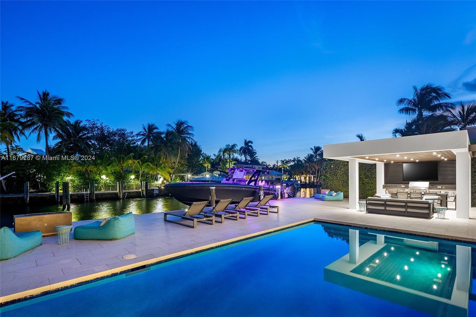 This image showcases a luxurious outdoor space featuring a stunning pool with crystal-clear water reflecting the surrounding architecture. The area includes modern lounge chairs, a boat docked nearby, and an outdoor kitchen/entertainment area with a large screen TV. The overall impression is one of high-end relaxation and entertainment, perfect for a waterfront property.