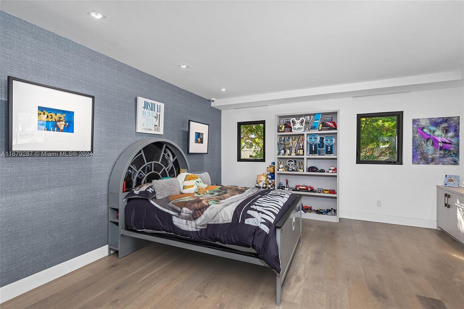This is a well-decorated bedroom featuring a unique bed with a Star Wars theme, complemented by a built-in shelving unit displaying trophies and collectibles. The room is illuminated by natural light from two windows, and the hardwood flooring adds warmth to the space. The walls are painted in a combination of blue and white, creating a playful yet sophisticated atmosphere.