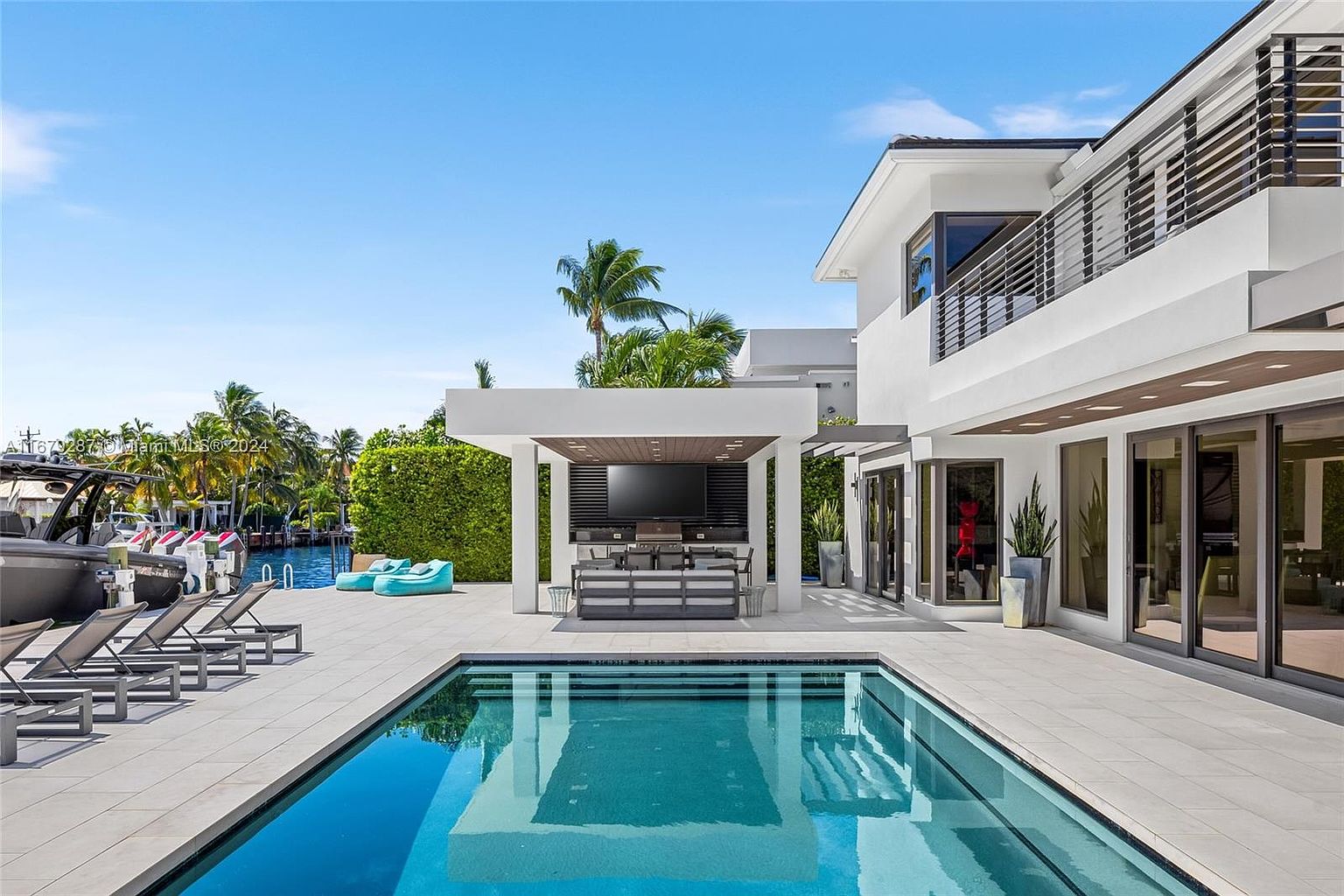This image showcases a luxurious backyard with a rectangular swimming pool as the focal point. The pool is surrounded by light-colored tiling and modern lounge chairs. A covered outdoor living area with a large screen TV and seating is adjacent to the pool, and the contemporary architecture of the house is visible in the background, featuring large windows and a balcony. The perspective is from the pool level, capturing the house and outdoor amenities.