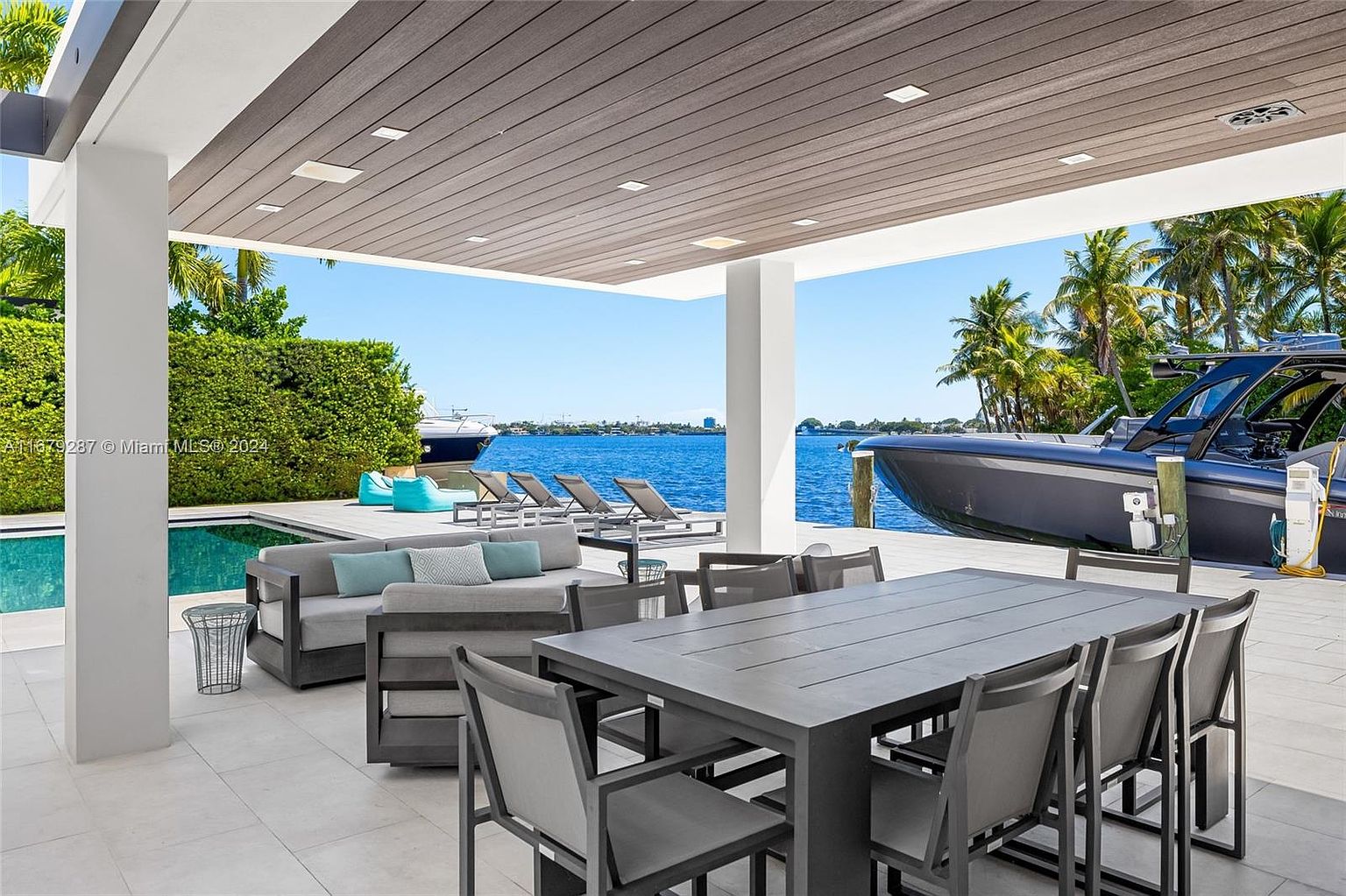 This image showcases a luxurious outdoor patio area with a modern aesthetic. The space features a large dining table with seating, a comfortable lounge area with a sofa and throw pillows, and several lounge chairs overlooking the water. A sleek boat is docked nearby, and the patio is covered by a stylish roof with recessed lighting, creating an inviting and upscale outdoor living space.