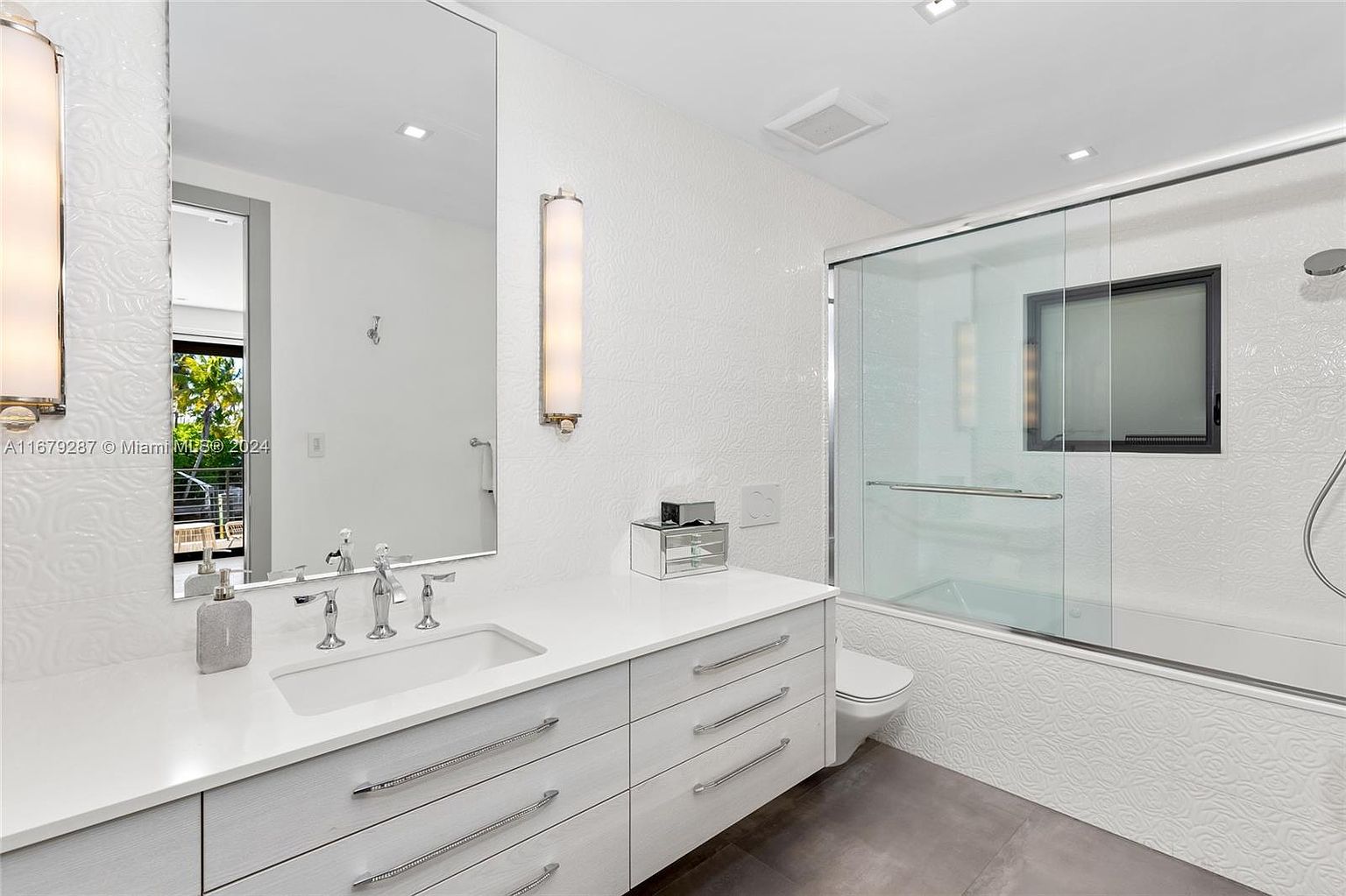 This is a well-lit primary bathroom featuring a modern vanity with a white countertop and light wood-tone drawers with sleek silver pulls. A large mirror reflects the outdoor light, and the textured white wall tiles add a touch of elegance. The shower area is enclosed with glass, and the overall design is clean and contemporary.