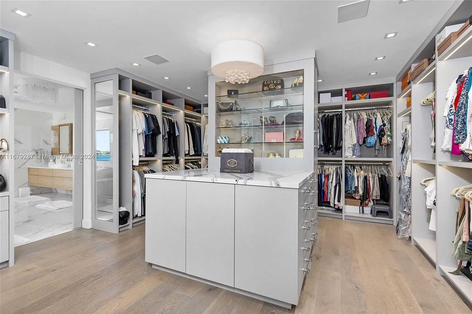 This is a spacious walk-in closet featuring custom shelving and hanging rods for organized storage. A central island with a marble countertop provides additional surface space, and a glass-enclosed display case showcases accessories. The closet is well-lit with recessed lighting and a decorative chandelier, creating a luxurious and functional space.