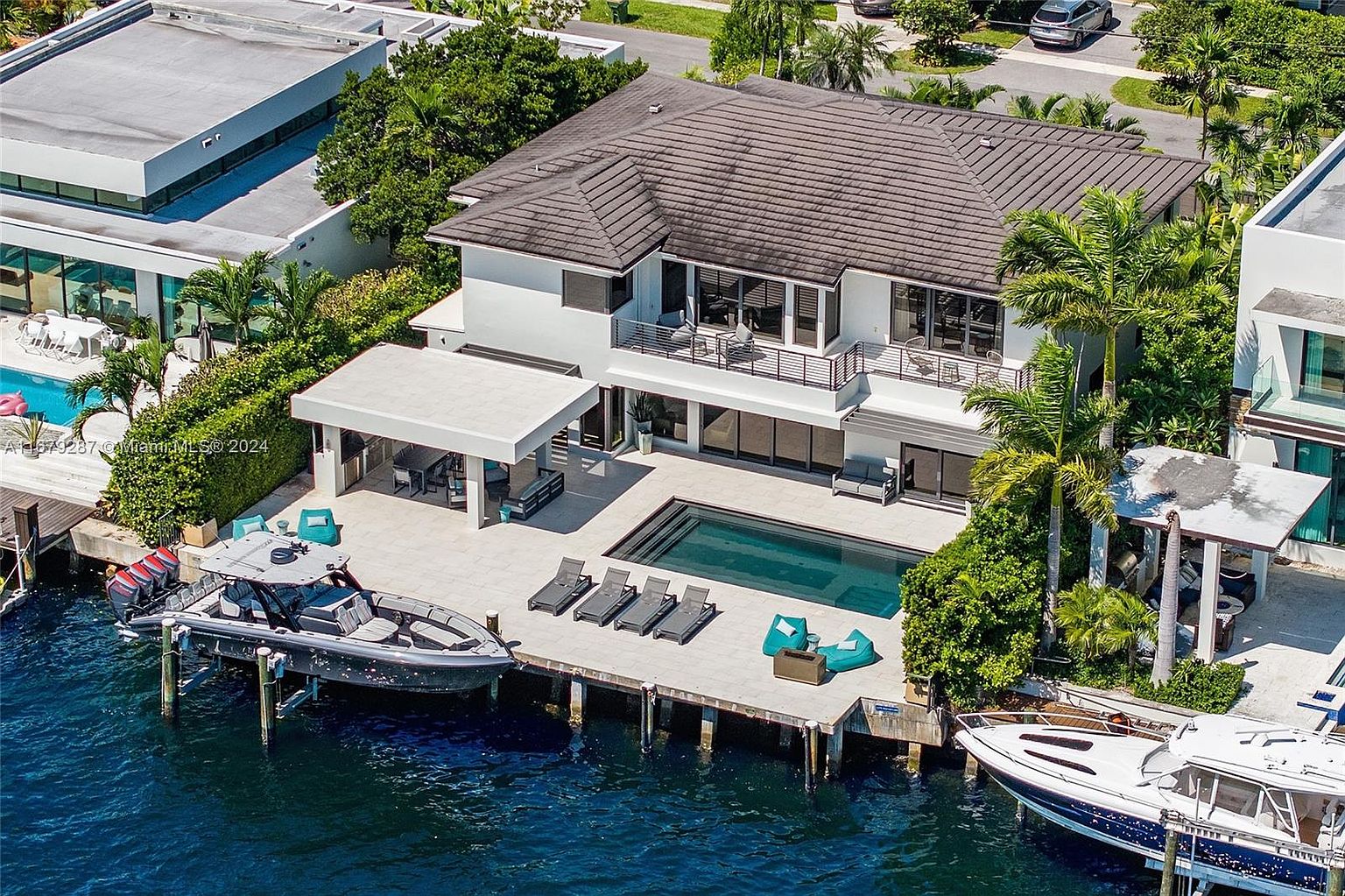 This aerial shot showcases a luxurious waterfront property with a modern architectural design. The house features a dark brown tile roof, white exterior walls, and multiple balconies overlooking a pool and dock. Two boats are moored at the dock, emphasizing the property's direct water access and upscale lifestyle.