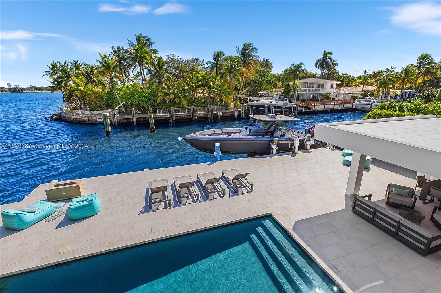 This image showcases a luxurious waterfront property featuring a modern pool and patio area. A sleek motorboat is docked alongside the property, emphasizing the waterfront access. The scene includes stylish outdoor furniture, lush tropical landscaping, and a glimpse of the contemporary architecture of the house, creating an impression of upscale coastal living.