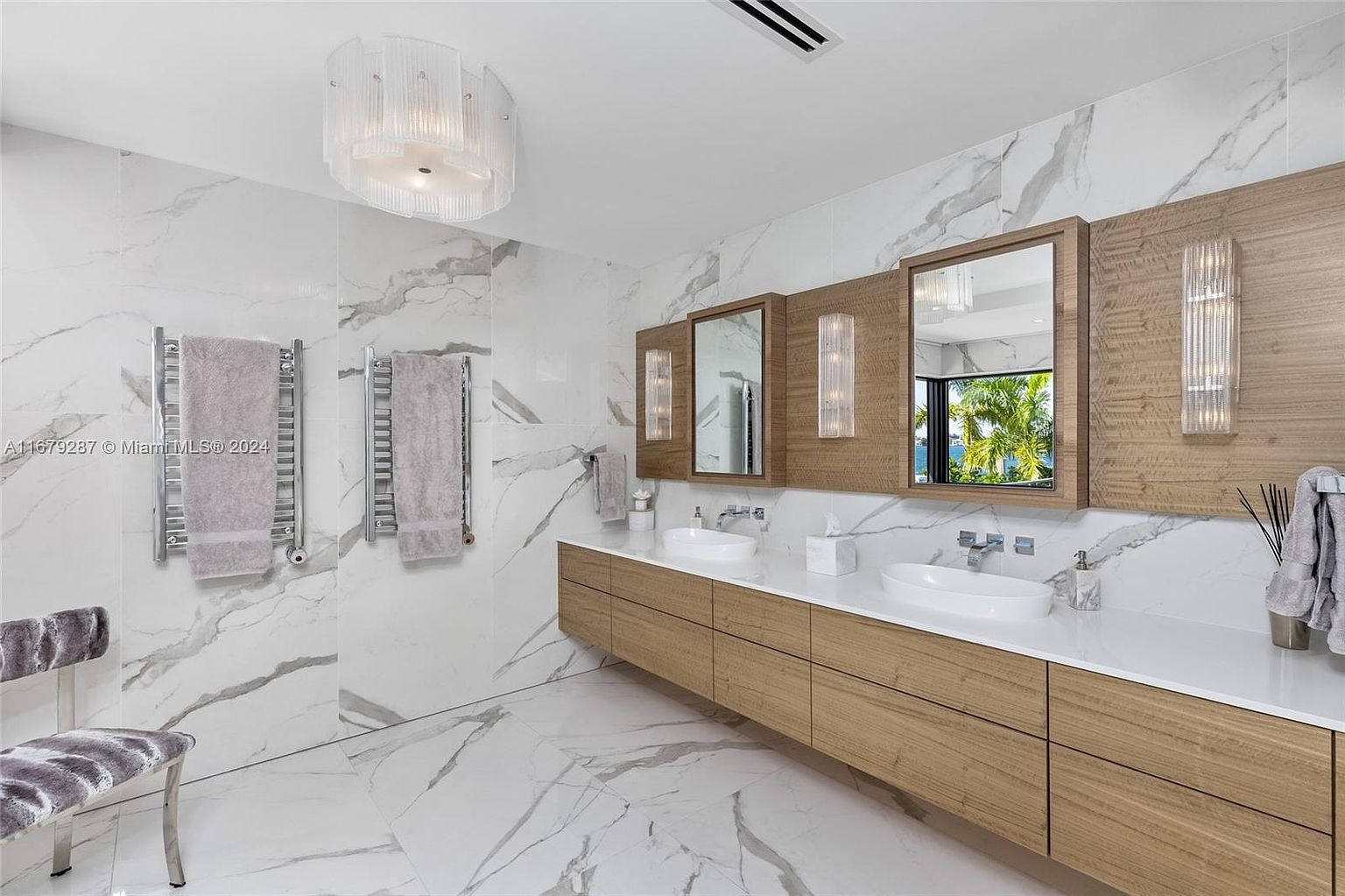 This is a luxurious primary bathroom featuring a double vanity with modern sinks and sleek wooden cabinetry. The walls and floors are covered in elegant marble, and the room is illuminated by a stylish chandelier and sconces. The overall impression is one of sophistication and high-end design.